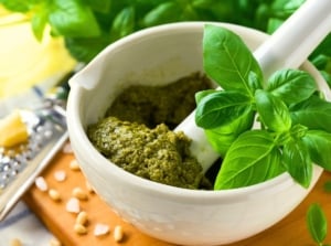 On the table there is a white bowl full of green pestle and fresh glossy leaves of the best basil for pesto among the ingredients and kitchen utensils.