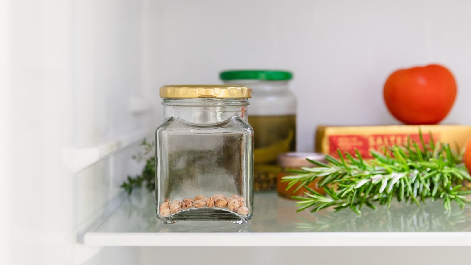 A small glass jar with a metal lid containing dry herbaceous material, placed on a white shelf next to rosemary and a red fruit.