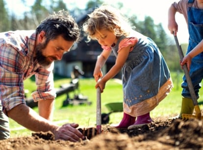 Father's Day gardening gifts. A father in a plaid shirt checks the soil in a bed that his two children, a girl and a boy, are raking in a sunny garden.