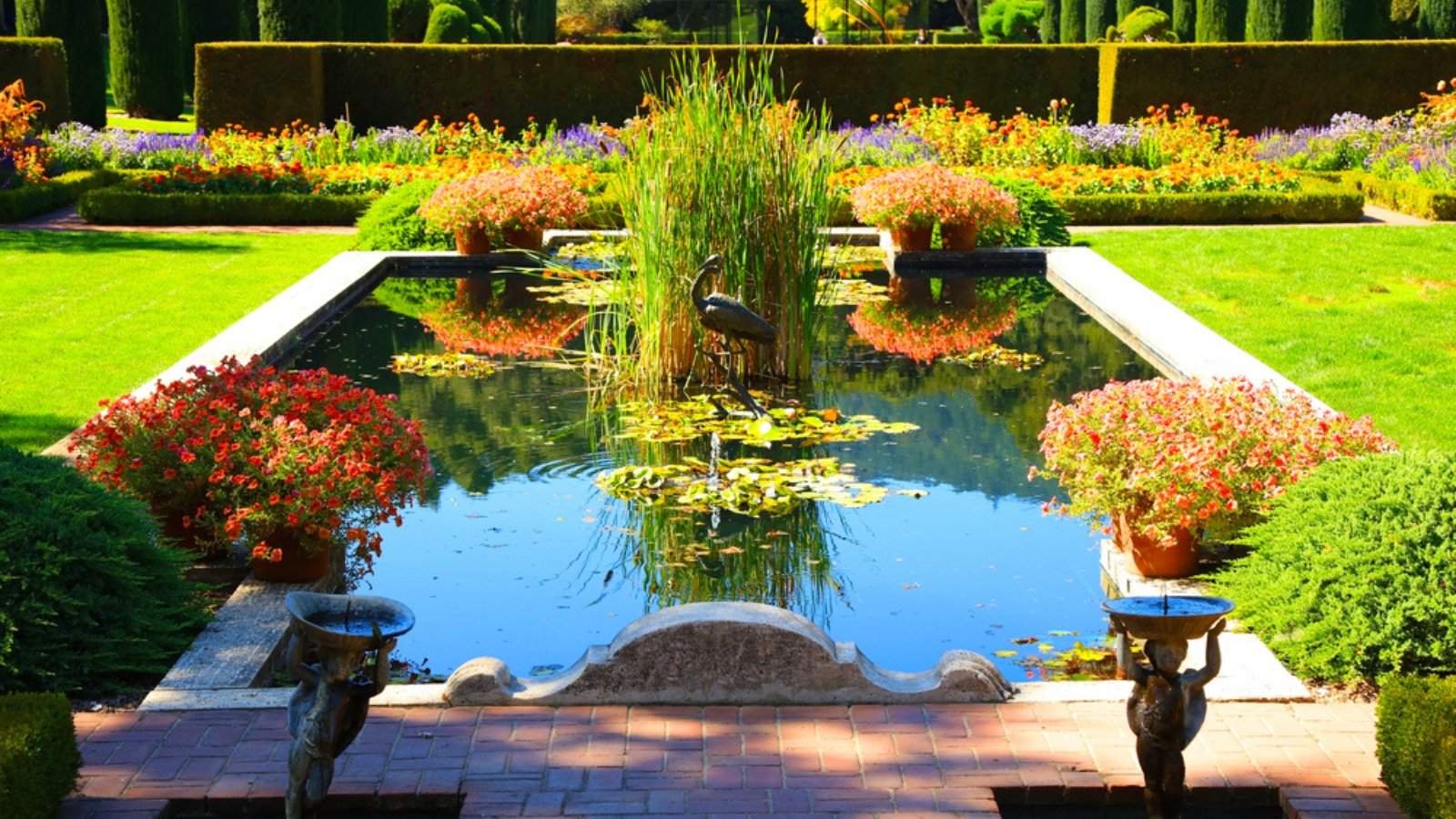 View of a sunlit garden with a reflecting pool surrounded by an array of blooming plants in elegant containers and meticulously groomed box bushes.