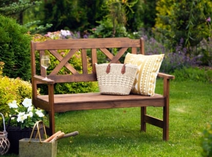 Upgraded backyard landscaping, rustic wooden bench with throw pillow, wicker bag and wine glass stands in the middle of green lawn surrounded by various bushes, trees and flowering plants
